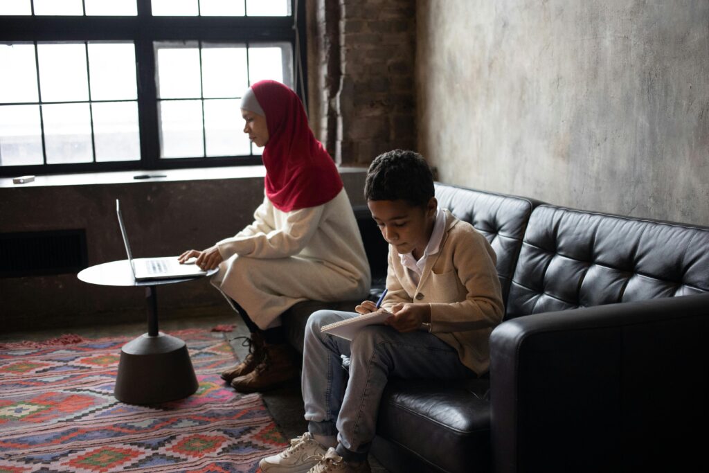 Side view of concentrated son preparing project for school while sitting on soft couch together with Arabian mother surfing laptop