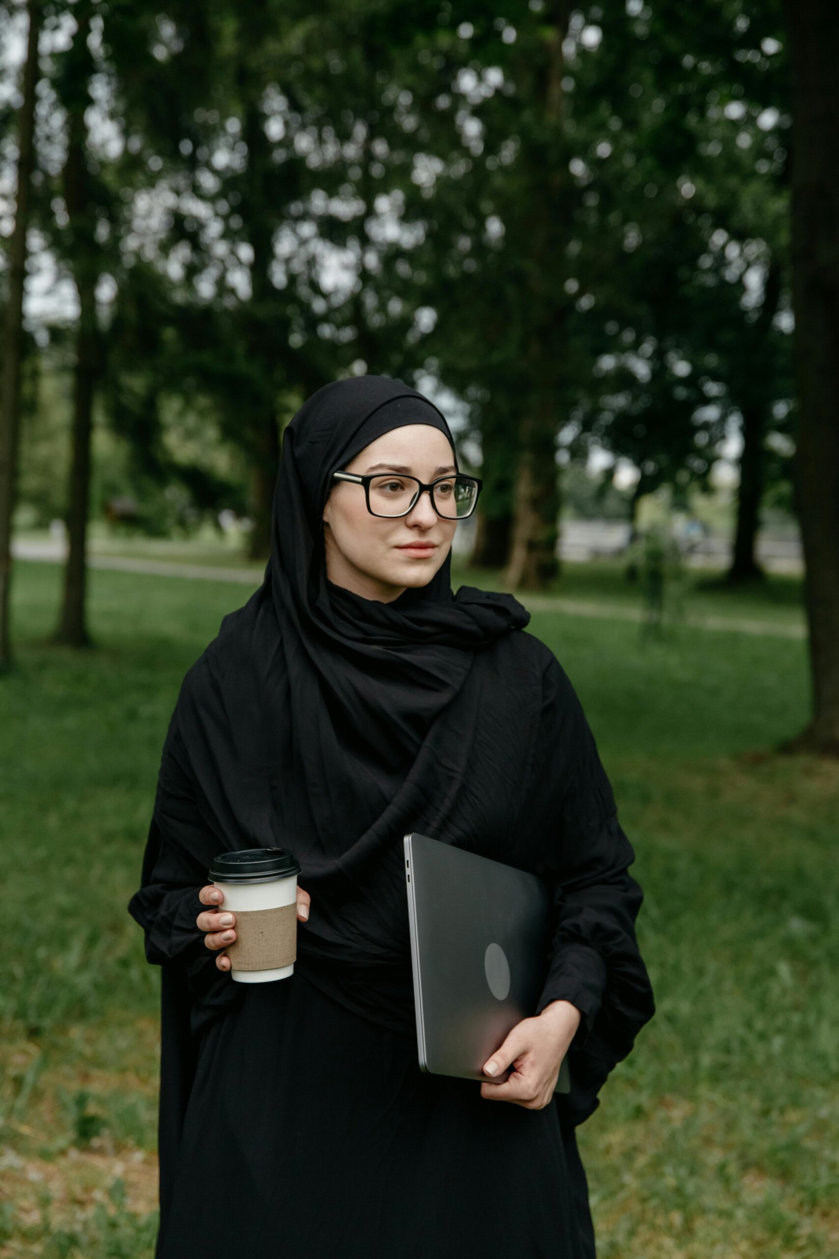 A young woman wearing a hijab and glasses holds a coffee cup and laptop in a serene park setting.
