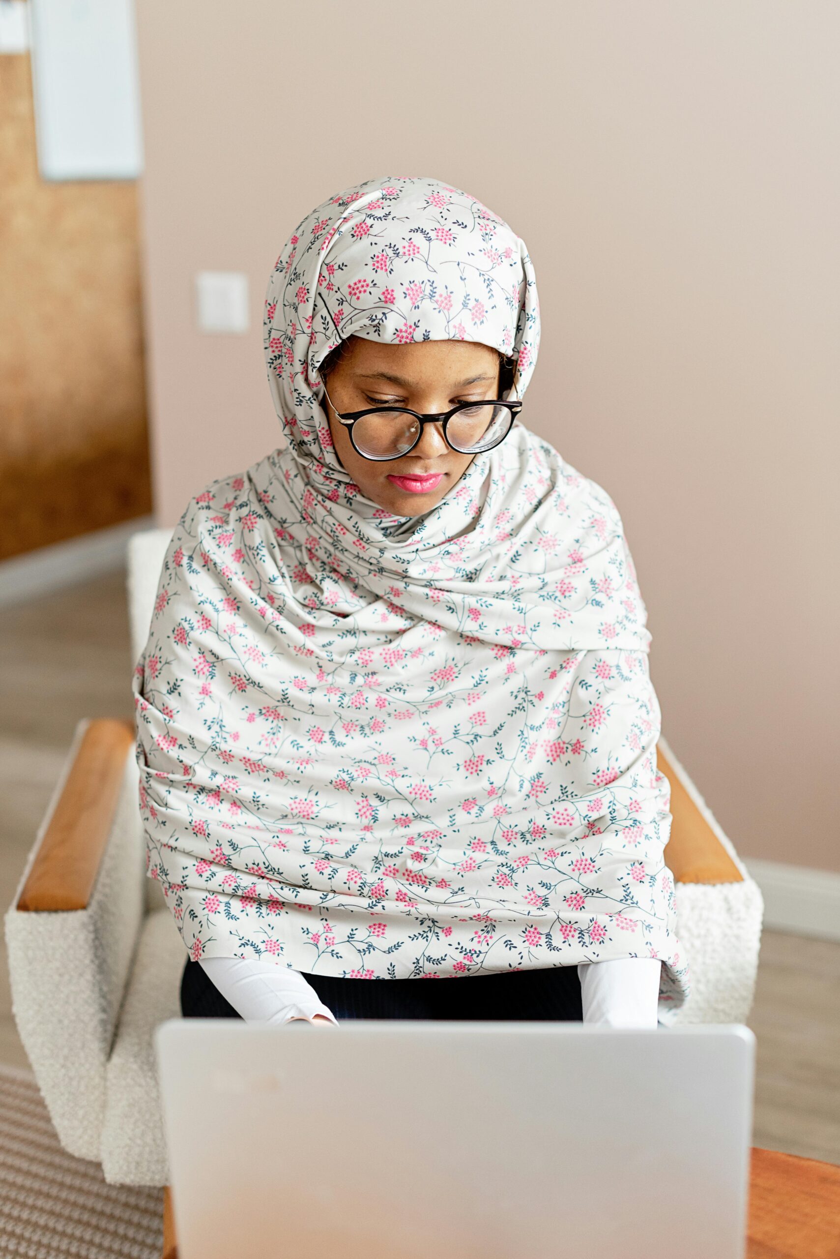 A Muslim woman with eyeglasses working on a laptop in a modern office setting.